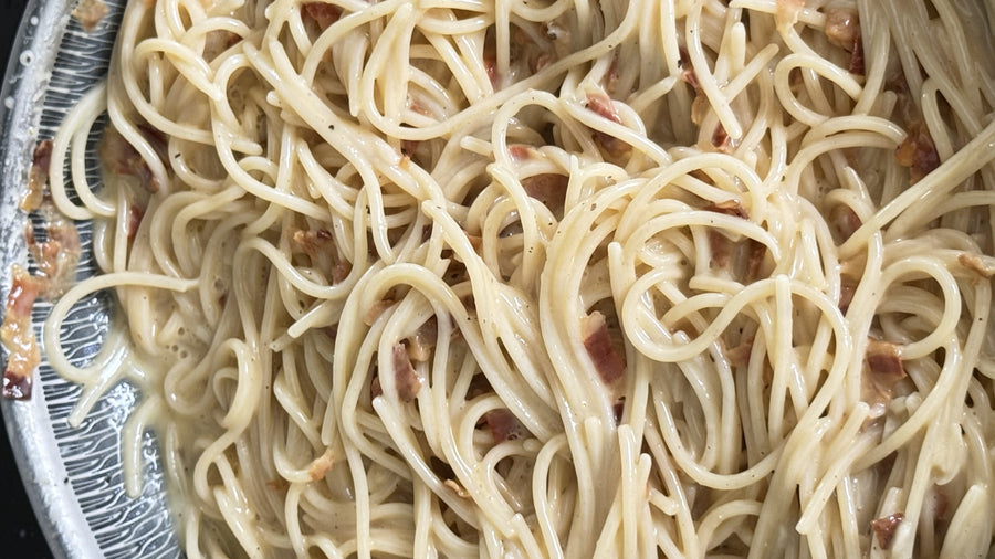 Spaghetti carbonara in a pan, with spaghetti coated in a creamy egg and cheese sauce, crispy pieces of cured pork and flecks of black pepper.